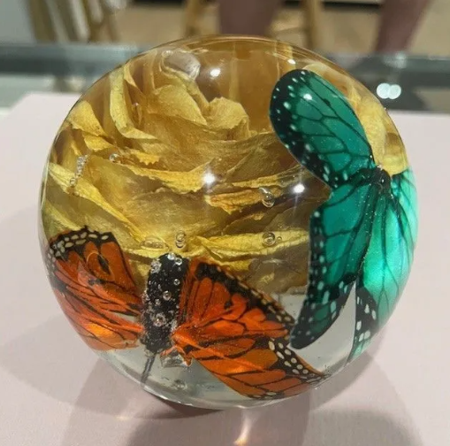 A clear sphere with preserved butterflies and yellow roses. Orange and teal wings stand out.