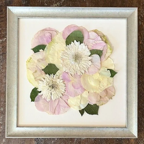 Framed artwork of dried flowers in soft pastel hues; silver frame on wood surface.