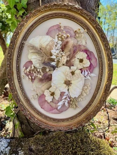 Oval framed dried flower arrangement in shades of white, pink, and brown, displayed outdoors.