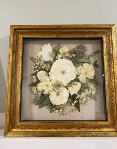 Framed pressed flower bouquet with white blooms, gold frame on a white surface.