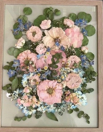 Dried flowers framed; pink and blue blooms, green leaves, light wood frame.