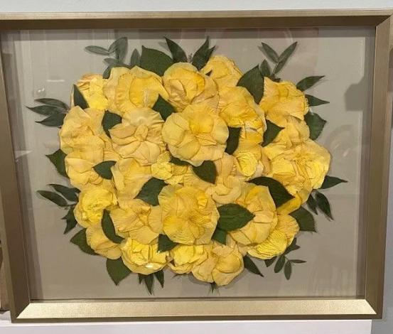 Yellow flower arrangement in a clear framed display, surrounded by green leaves on a light background.