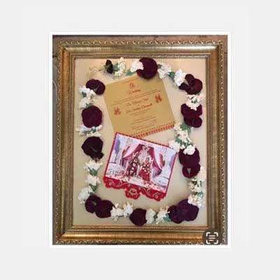 Framed wedding invitation and photo surrounded by maroon petals and white flowers.