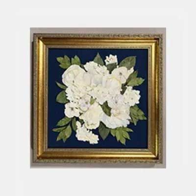 Framed pressed flower art, white flowers and green leaves on dark blue background in ornate gold frame.