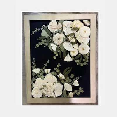 Framed floral arrangement. White pressed flowers and green sprigs on a dark background. Silver frame.