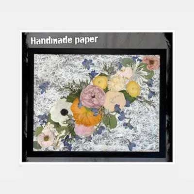 Handmade paper artwork: pressed flowers in a black frame, on a textured white background with scattered blue flowers.