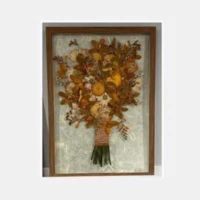 A framed dried flower bouquet art piece with an orange and yellow color palette, on a white textured background.