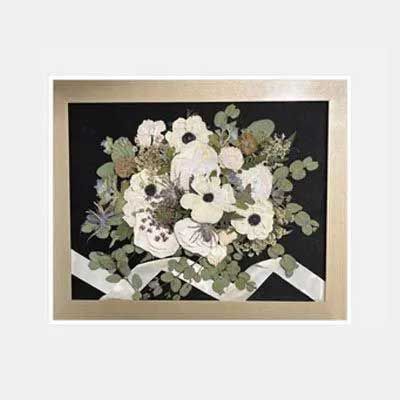 Framed pressed flower bouquet with white blooms, greenery, and ribbon on a black background.