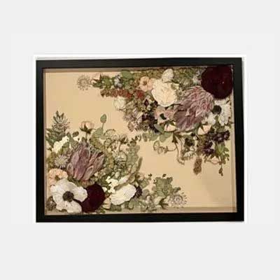 Framed floral arrangement featuring dried flowers in cream, burgundy, white, and green on a tan background.
