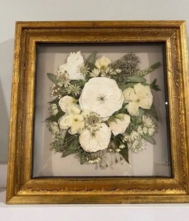 Framed dried flower arrangement with gold frame. White and cream colored flowers on a beige background.