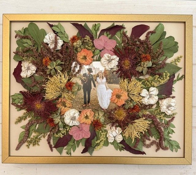 Pressed-flower wedding scene in a gold frame with colorful petals and leaves on a beige background.