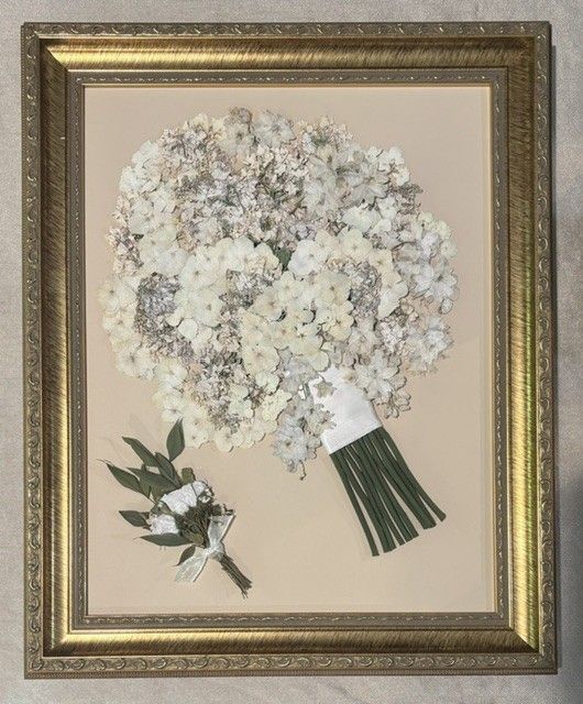 Framed pressed white flower bouquet with green stems and a small matching sprig on a beige background.