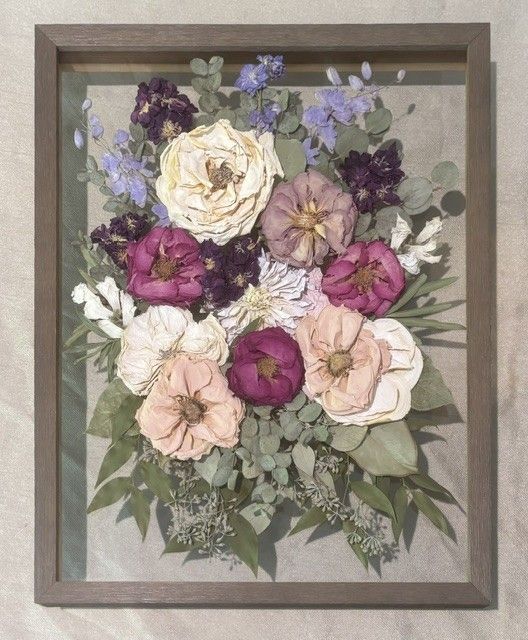 Framed pressed flower bouquet with cream, pink, purple, and green blooms on a gray background.