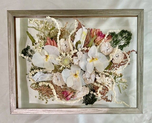 Framed pressed flower arrangement with white, pink, and green blossoms on a pale background.