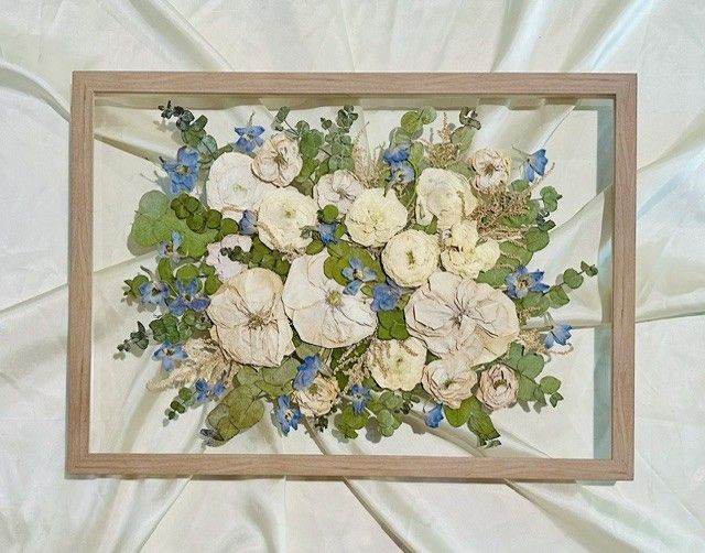Framed pressed-flower arrangement with white and blue blossoms on a white fabric background.