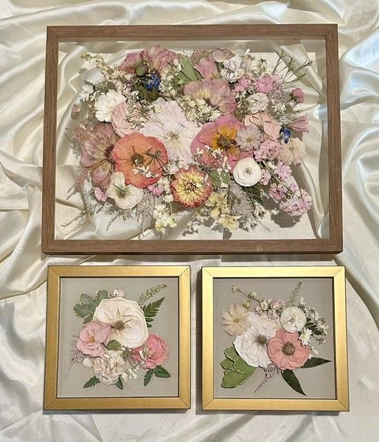 Three framed pressed-flower artworks on a white surface, in beige and gold frames with pink, white, and yellow blooms.