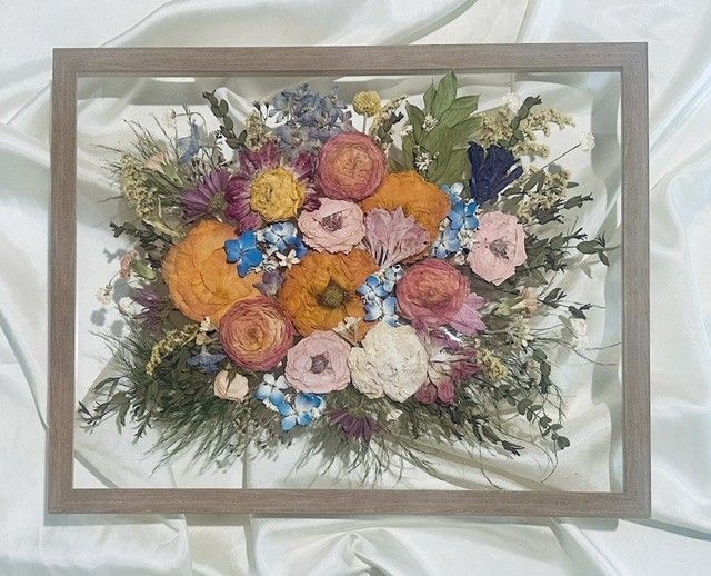 Framed bouquet of dried flowers in pink, orange, yellow, blue, and white on a light fabric background.