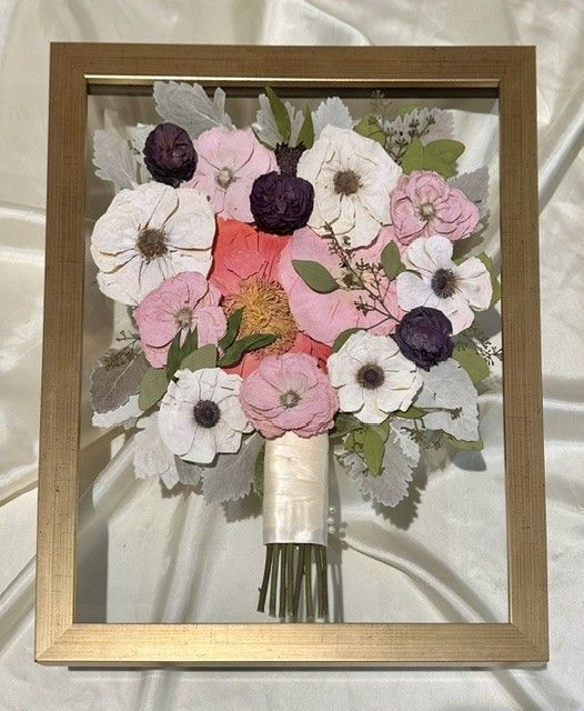Framed bouquet of pink, white, and purple flowers with green leaves on a beige background.