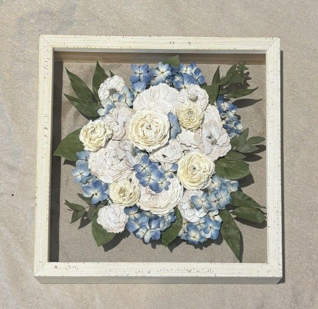 Framed floral arrangement with white and pale blue flowers and green leaves on a beige background.