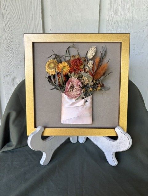 Framed dried flower bouquet in a gold frame on a white display stand against a gray backdrop.