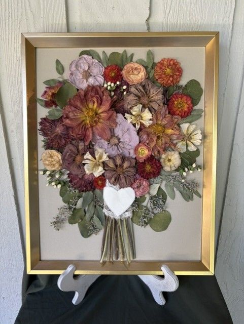 Framed pressed-flower bouquet with burgundy, pink, and green blooms in a gold frame on a stand.