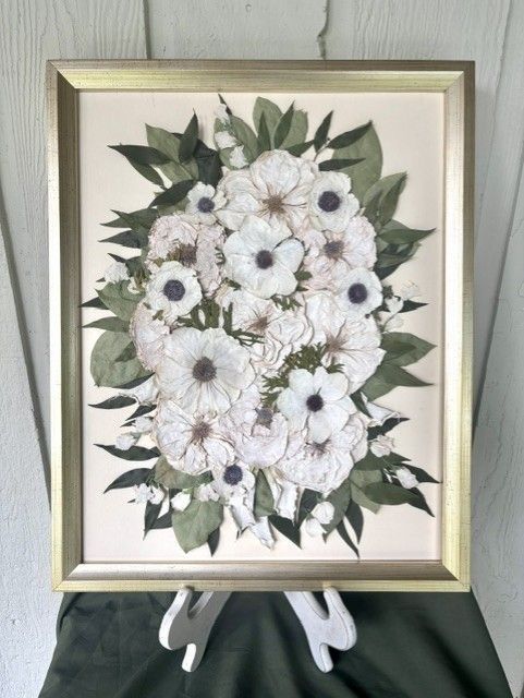 Framed floral arrangement with white flowers and green leaves on an easel.
