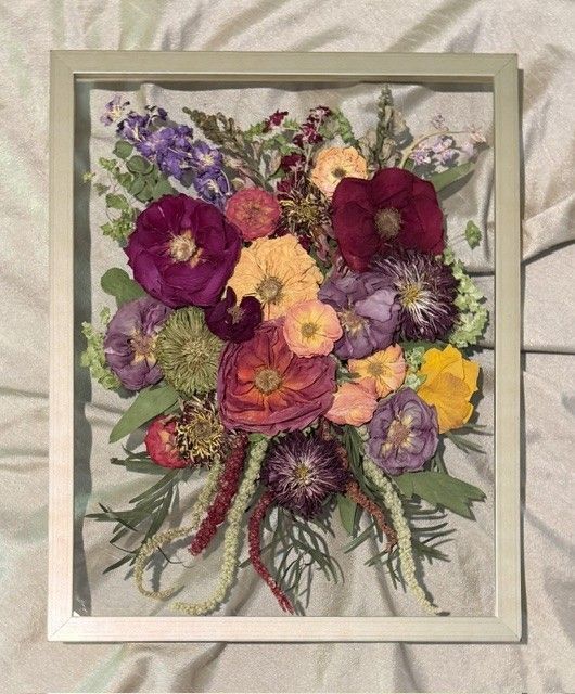 Framed bouquet of colorful pressed flowers with purple, pink, yellow, and green blooms on a light background.