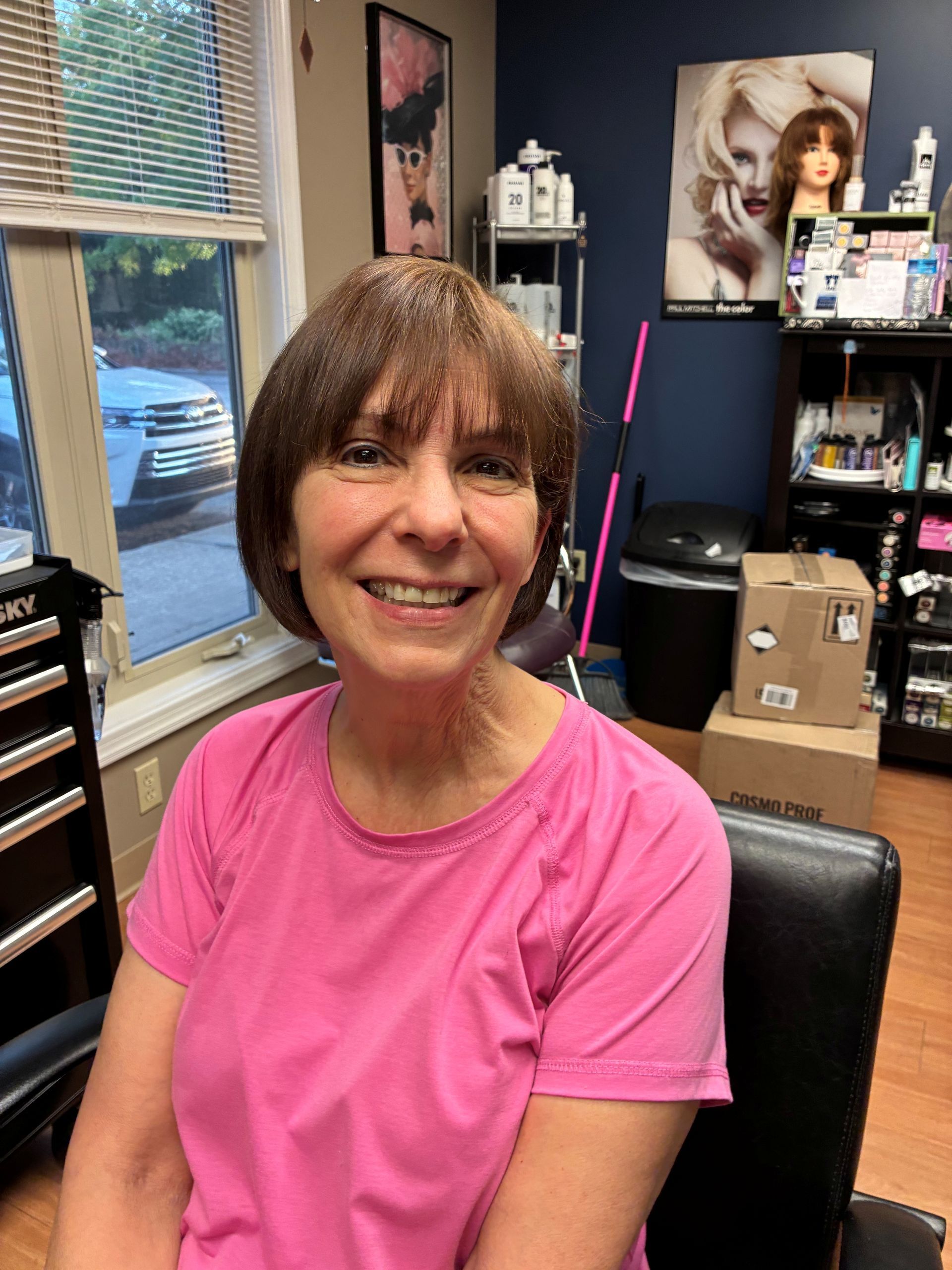 Smiling person in a bright pink shirt seated in an office with shelves and wall art behind them