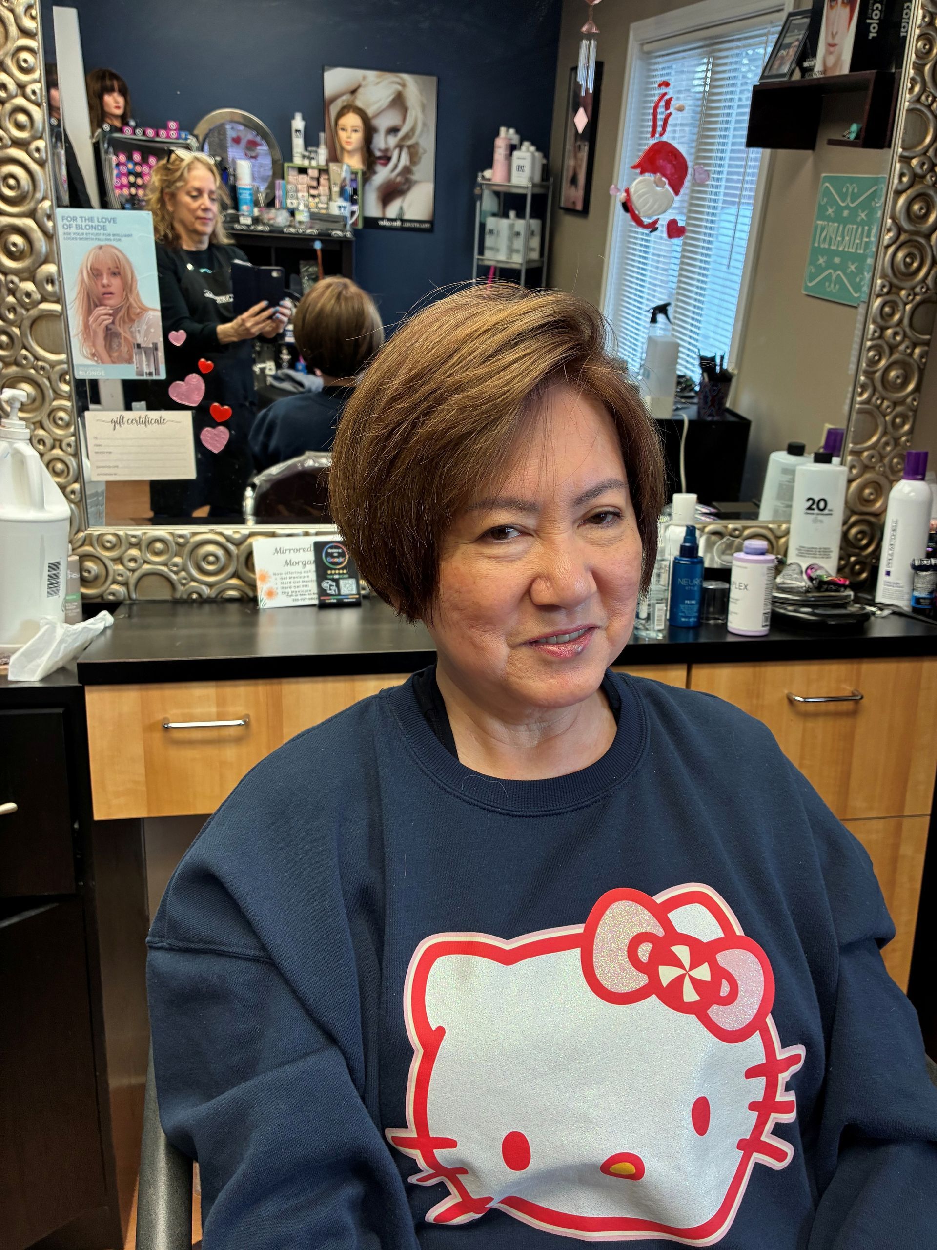 Woman smiling in a salon chair, wearing a navy Hello Kitty sweatshirt, with mirrors and hair products behind her
