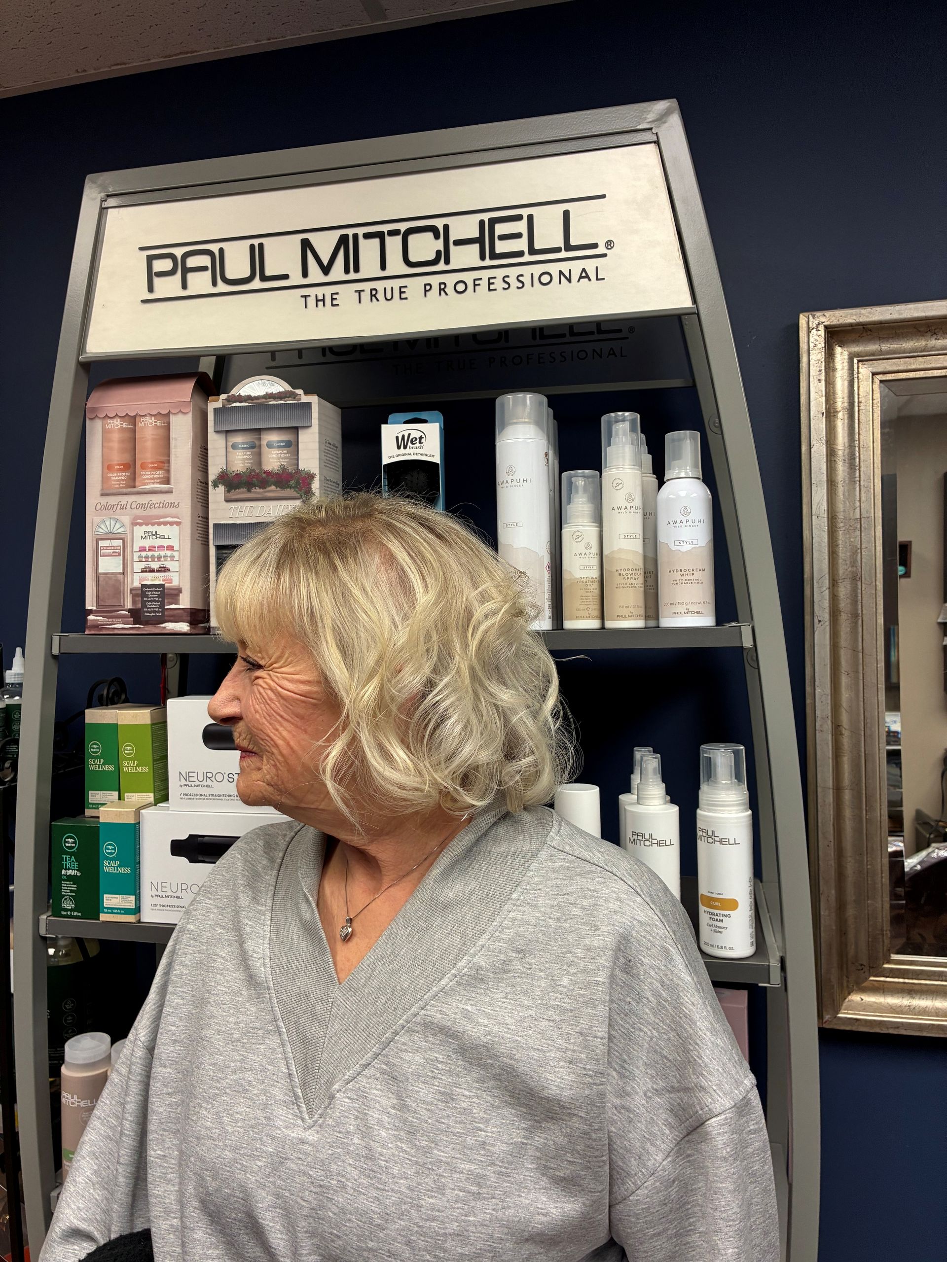 Woman with blonde hair standing in a salon aisle beside Paul Mitchell hair products.