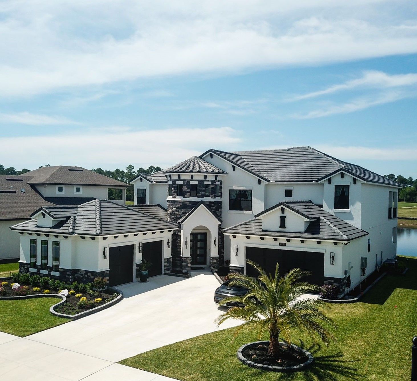 Large white Mediterranean-style house with dark roof, driveway, and palm tree in a sunny suburban neighborhood
