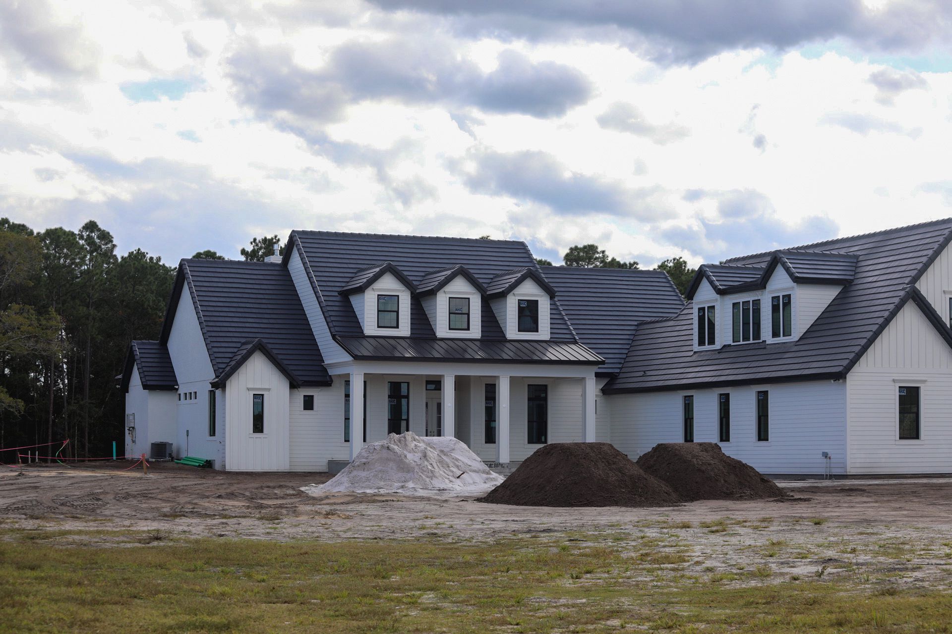 Large white house with a dark roof under construction, with dirt piles in the front yard under a cloudy sky