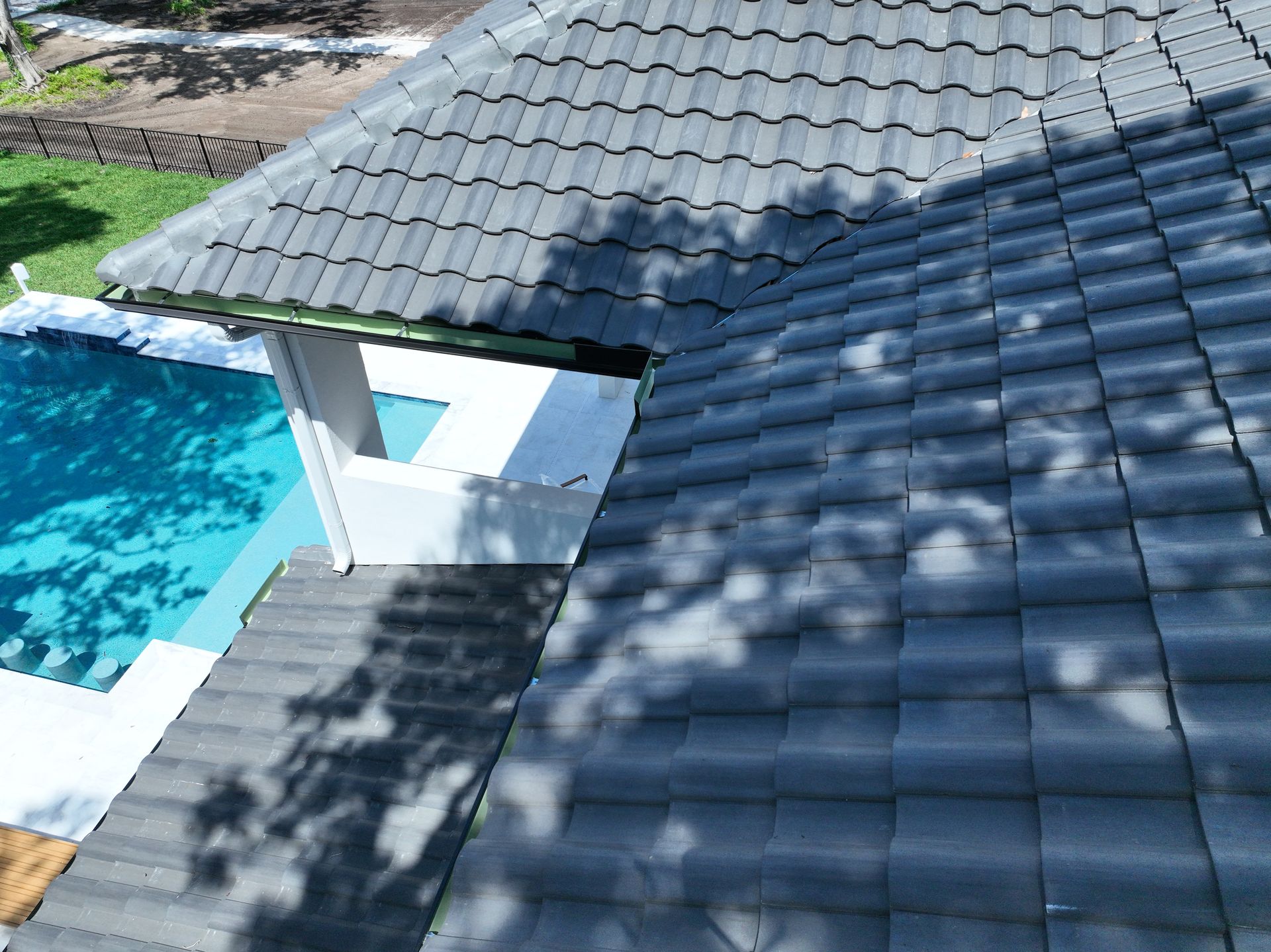 Dark gray tiled roof beside a blue swimming pool, with a small covered opening and tree shadows.
