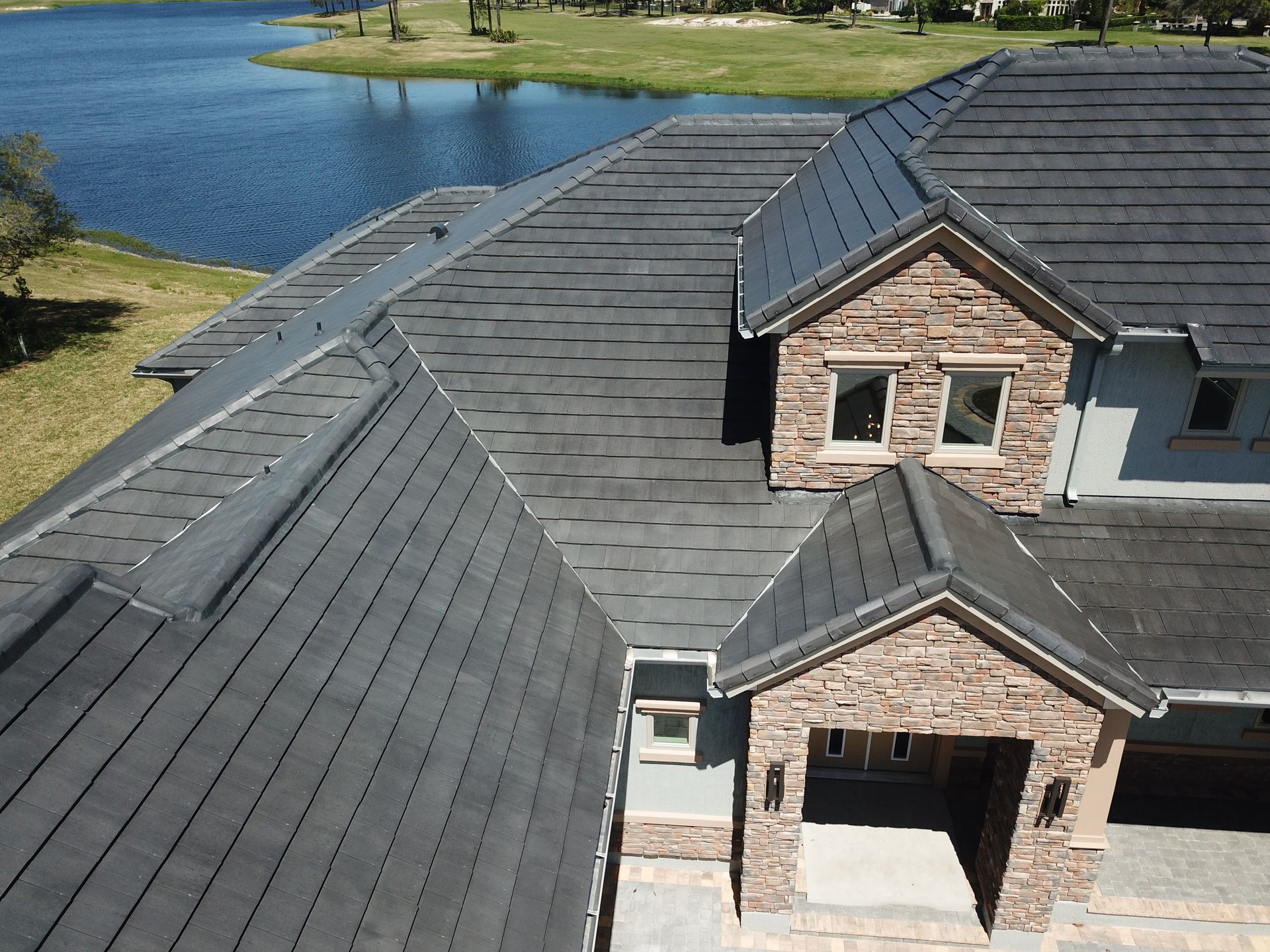 Aerial view of a modern house with dark gray shingled roof beside a pond and grassy yard.
