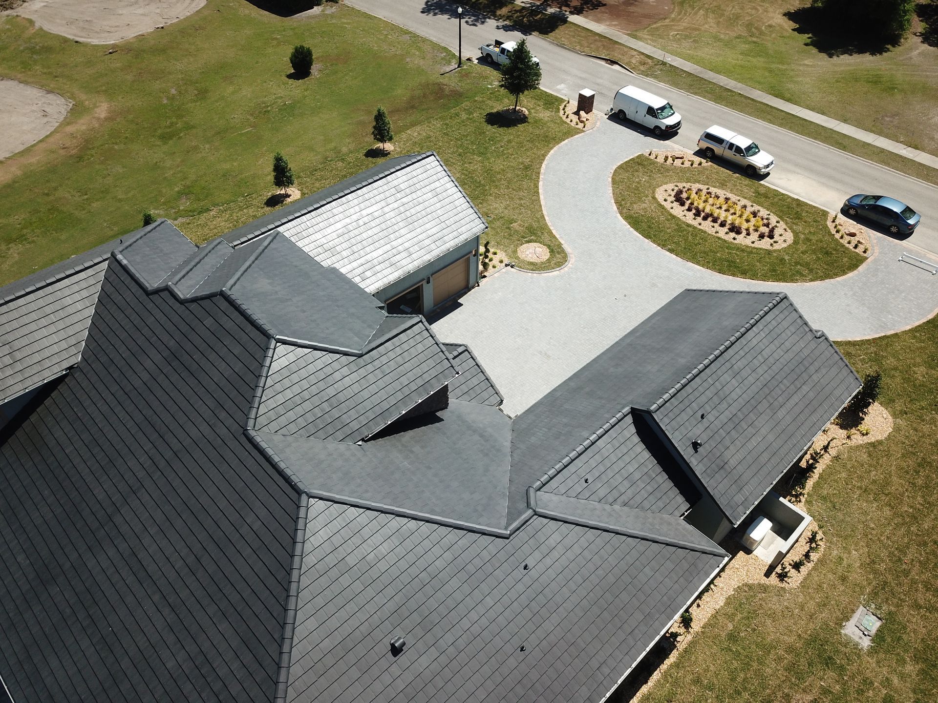 Aerial view of a house with gray roofs, driveway, and three parked vehicles on a green lawn.