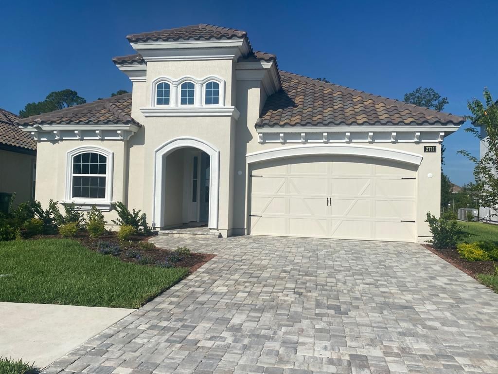 Two-story stucco house with a tiled roof, arched entry, and paver driveway under a clear blue sky