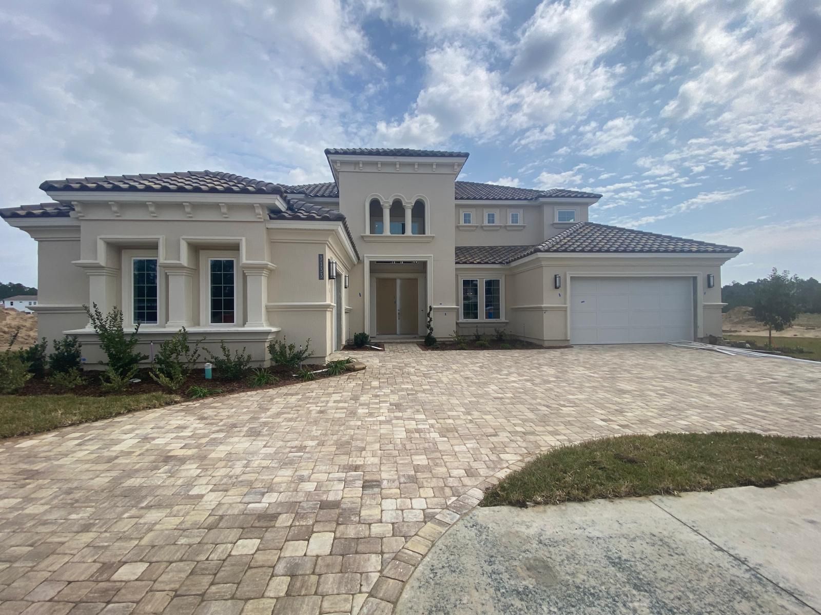 Large beige stucco house with a tiled roof, wide driveway, and attached garage under a cloudy sky