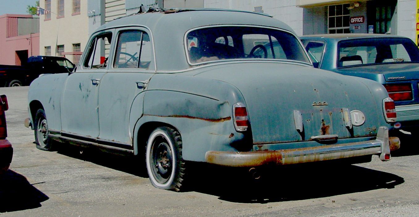 A blue old junk car with white tires is parked in a parking lot