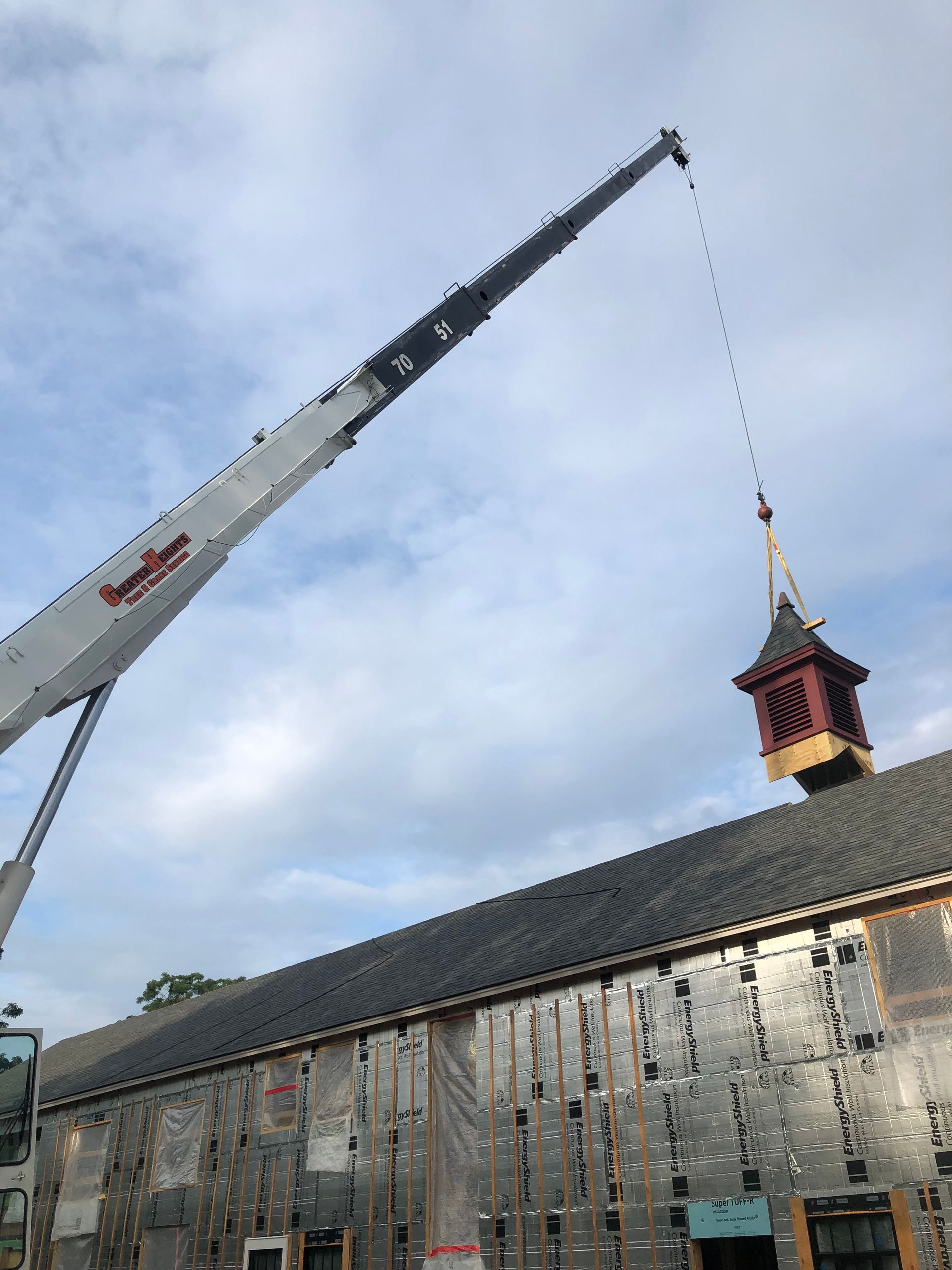A construction crane lifts a cupola onto the roof of a building covered in reflective silver insulation.