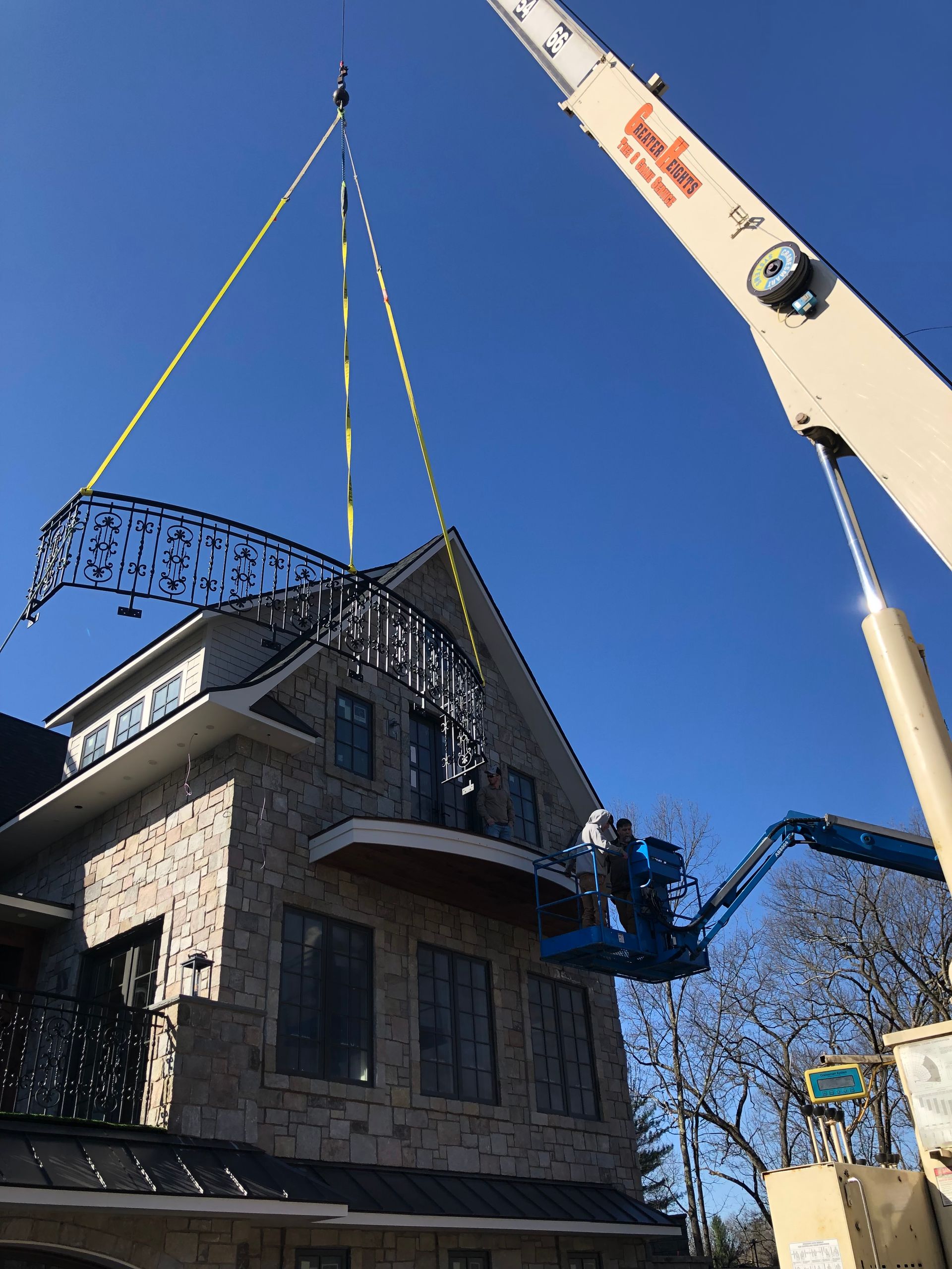 A crane lifts a decorative wrought-iron archway into place above a stone building's balcony under a clear blue sky.
