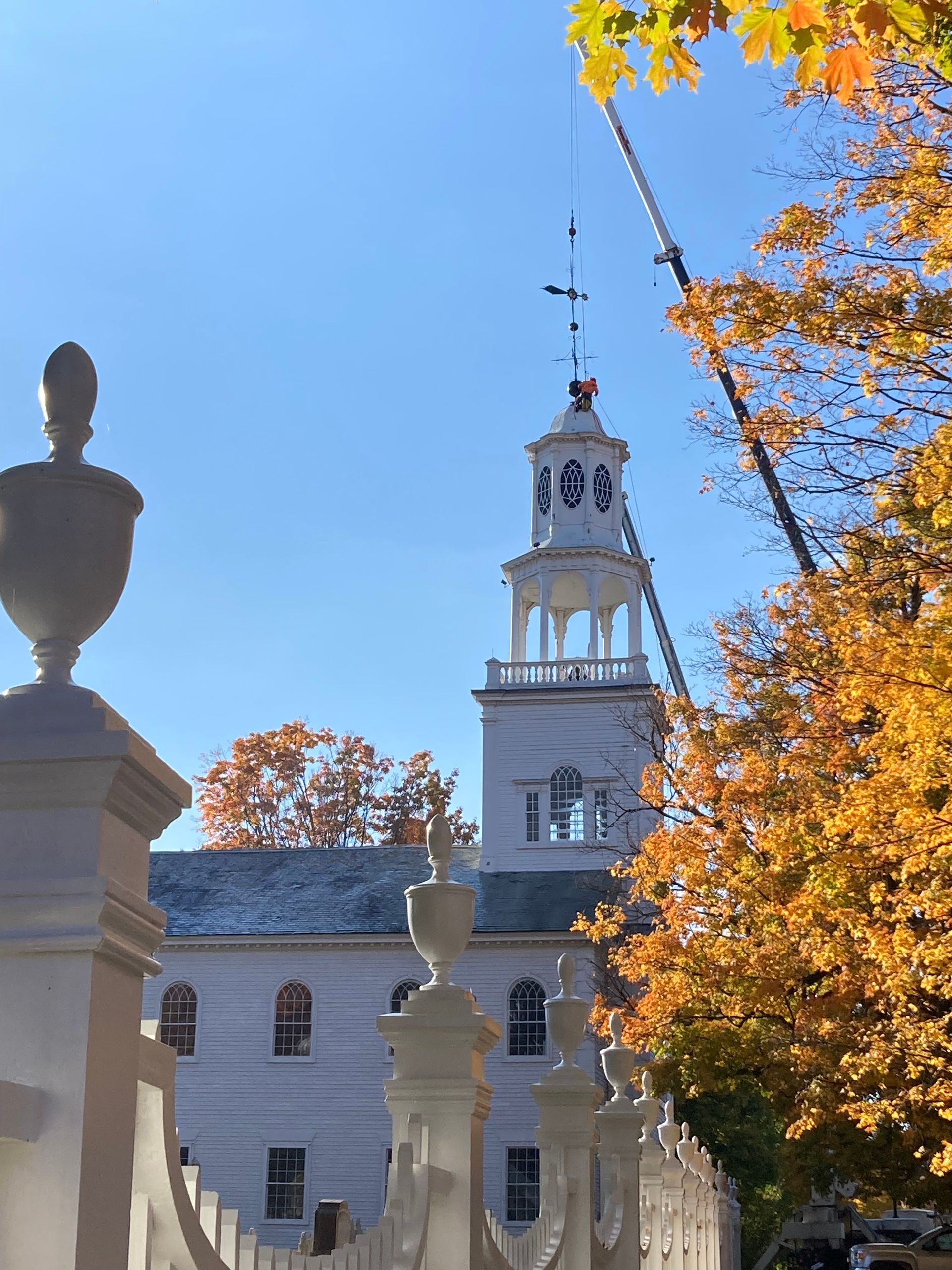 A crane works on the white steeple of a colonial-style church surrounded by autumn trees, viewed through an ornate fence.