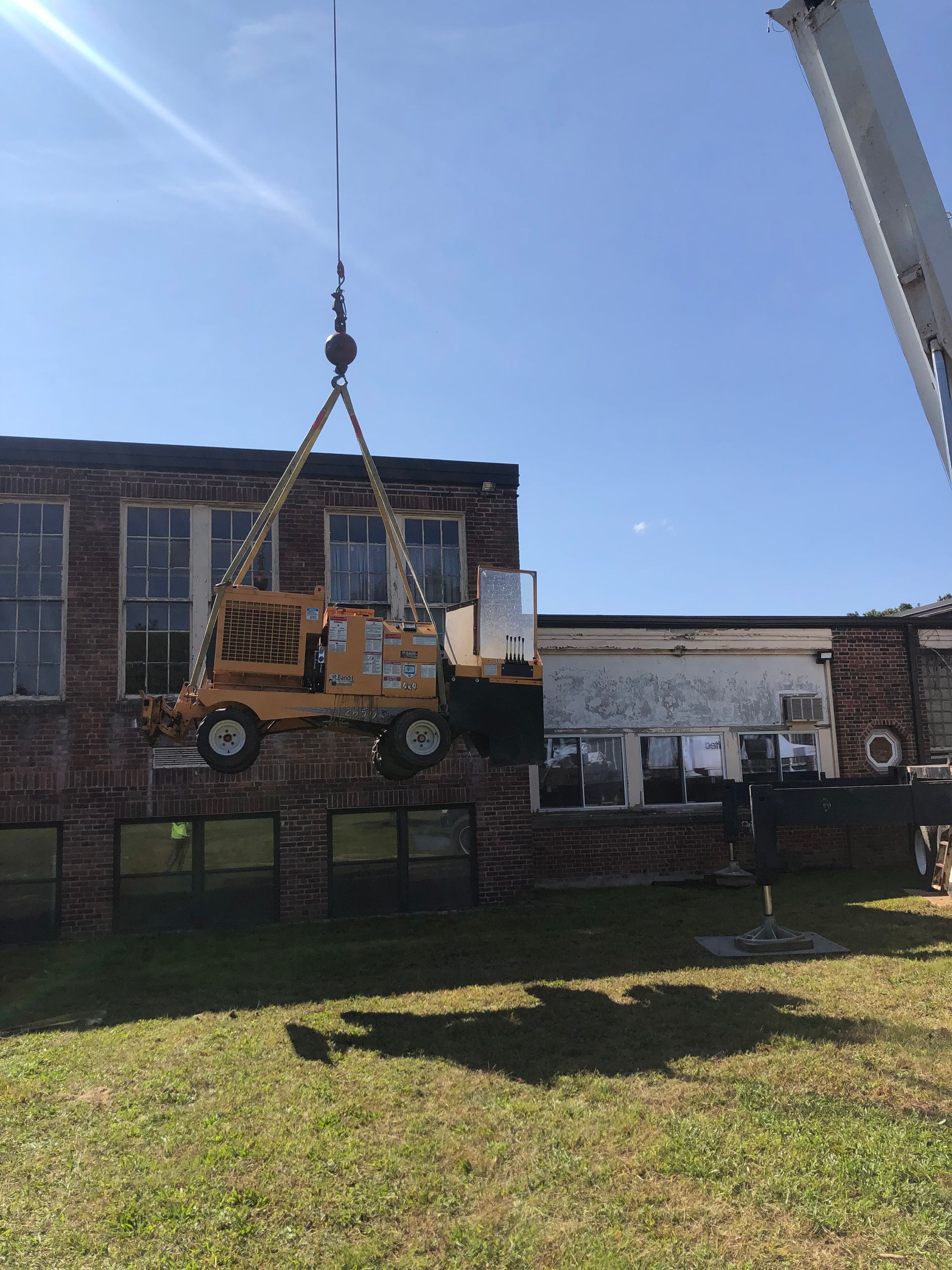 A construction crane lifts a yellow piece of heavy machinery in front of a brick building on a sunny day.