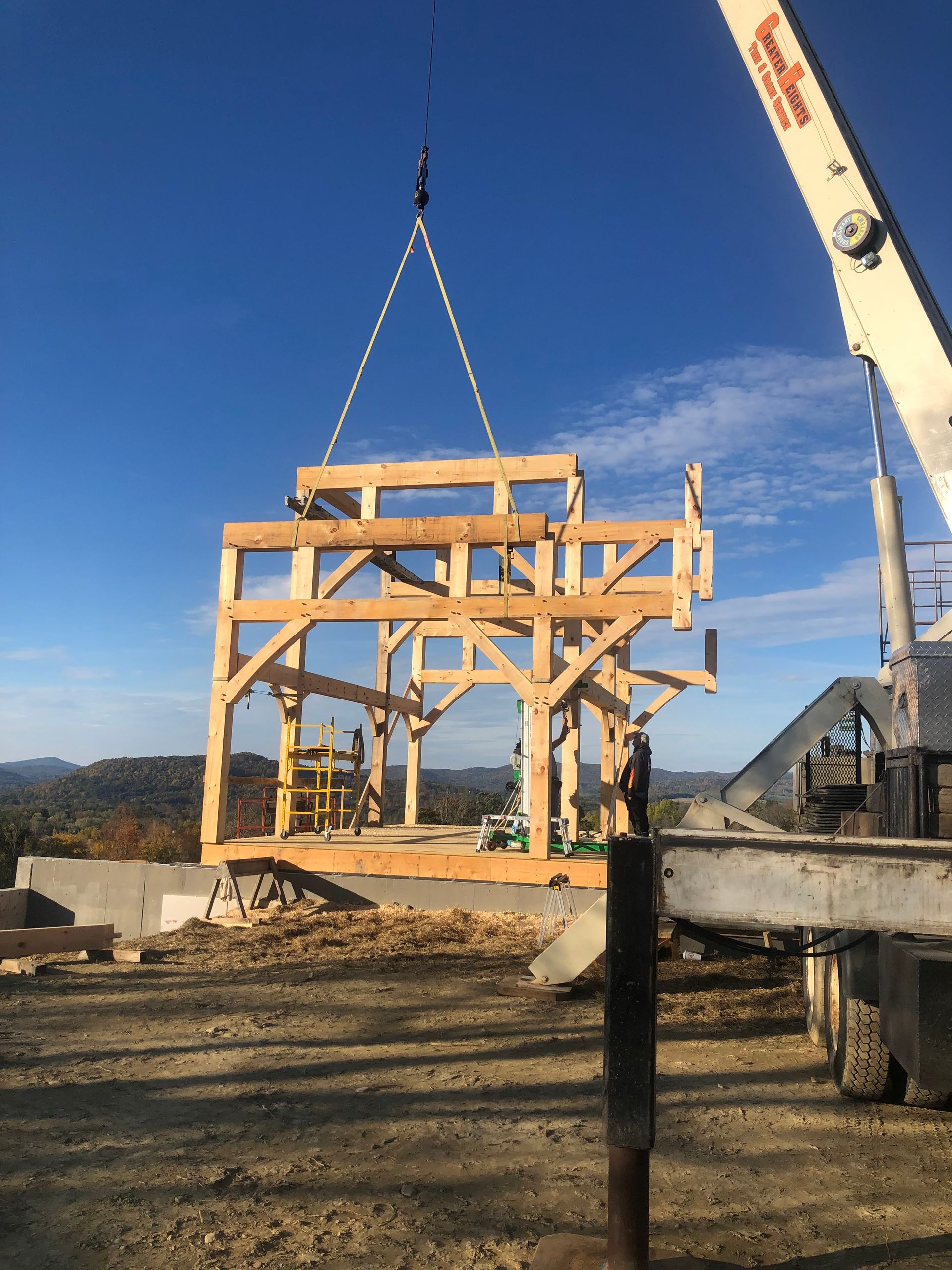 A crane suspends a timber-frame building skeleton above a concrete foundation on a sunny, rural construction site.