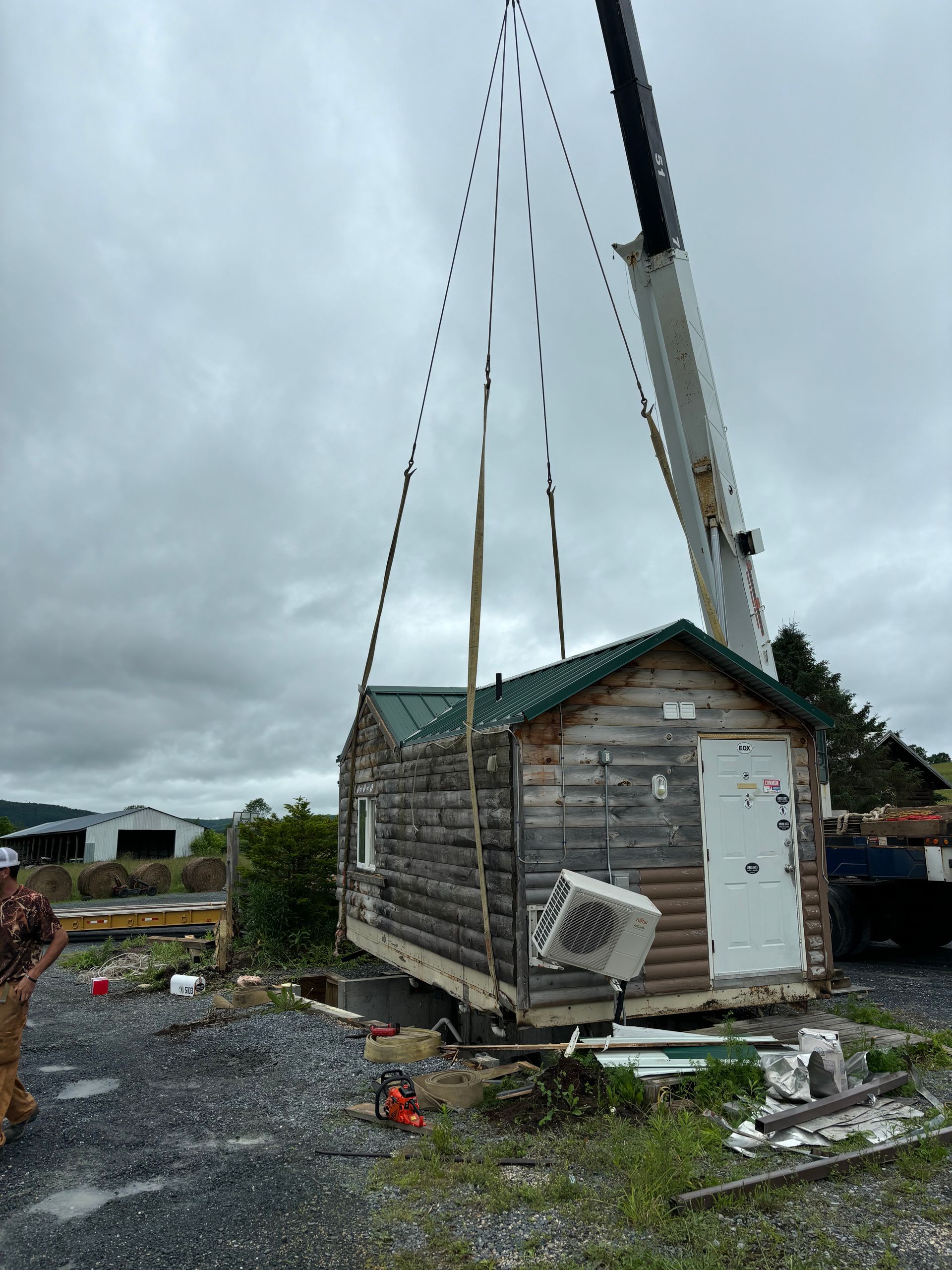 A crane lifts a small, wooden-sided cabin with a green metal roof into the air over a gravel construction site.
