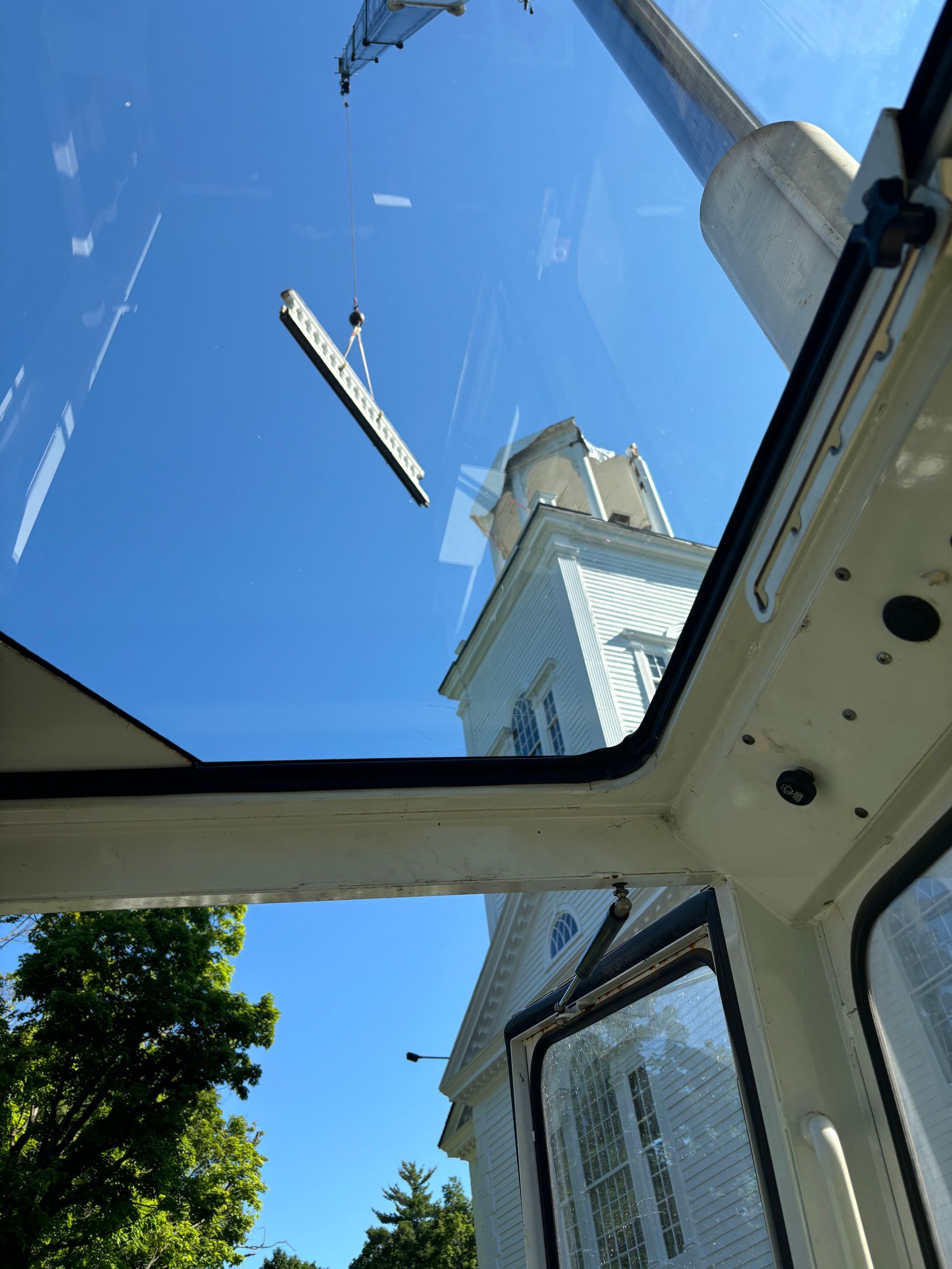 View from a crane operator's cab as a beam is lifted toward the steeple of a white church on a clear, sunny day.
