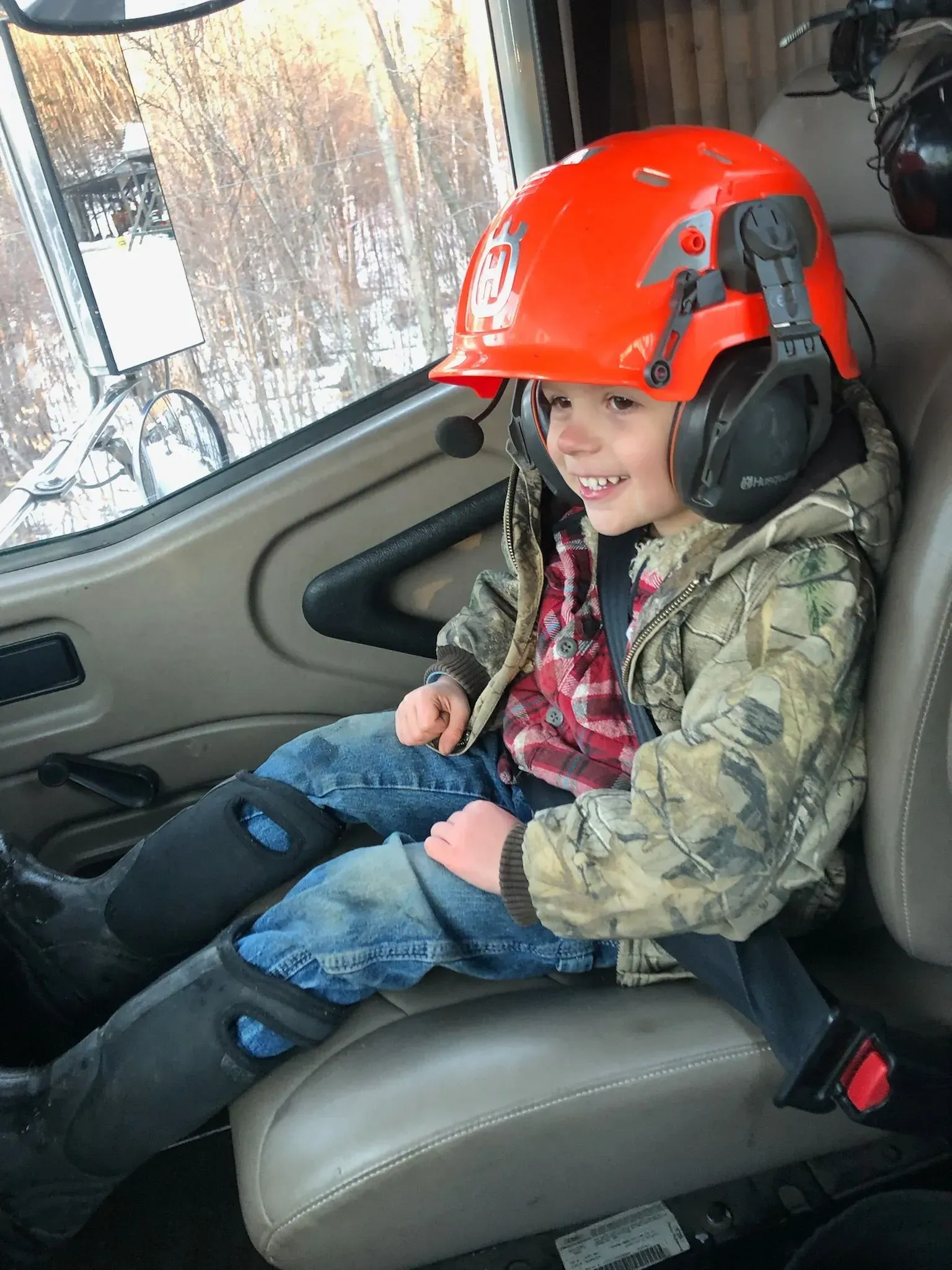 Child wearing a hard hat and earmuffs smiles while sitting in a large vehicle.