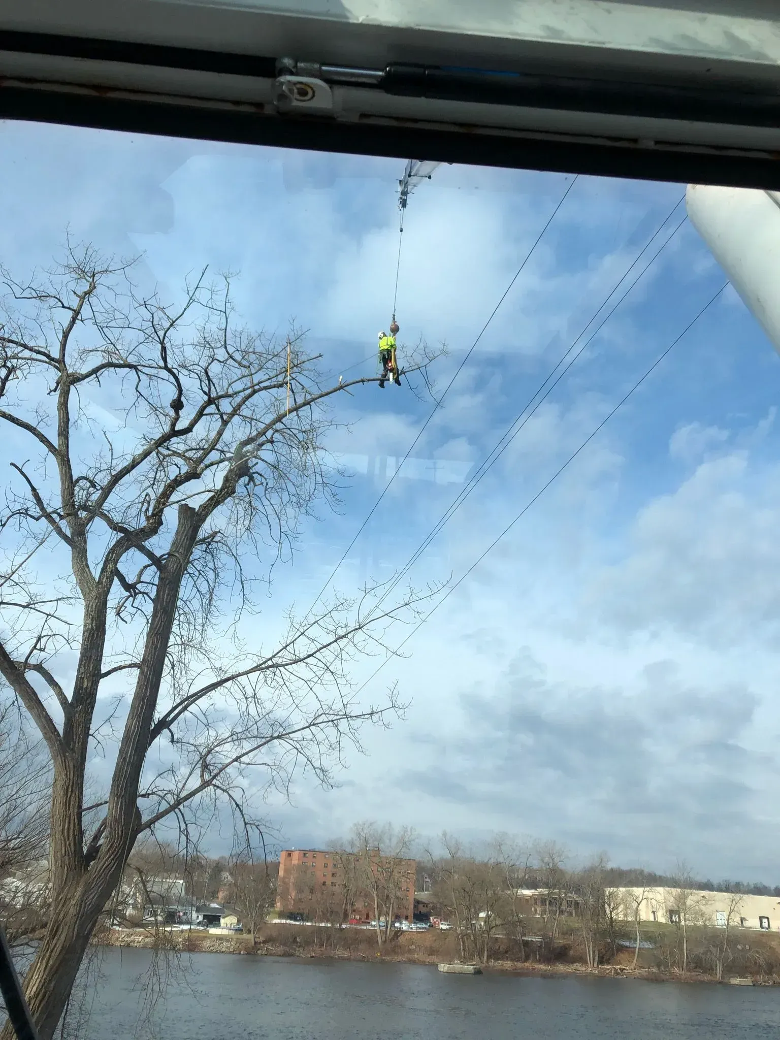 A utility worker in a high-visibility vest is suspended from power lines above a river with trees and buildings nearby.