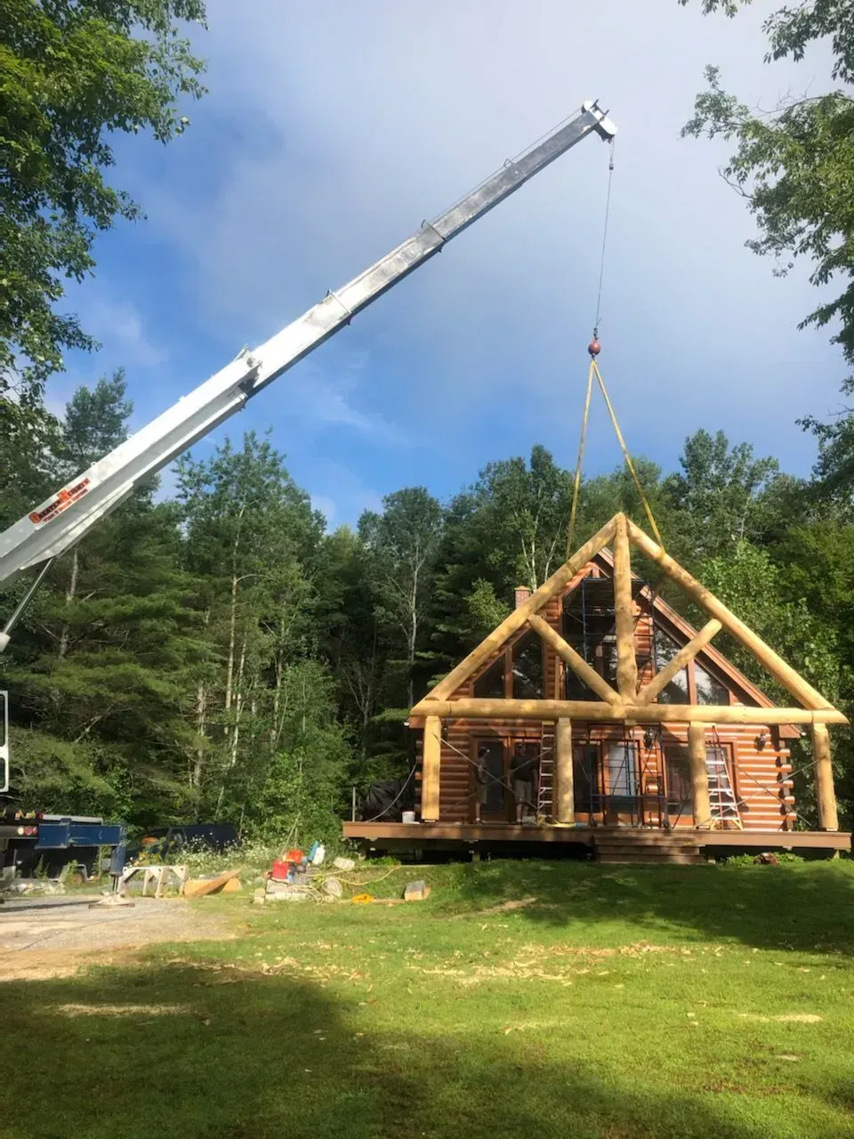 A crane lifts a large log frame gable onto a wooden log cabin in a grassy, wooded setting.