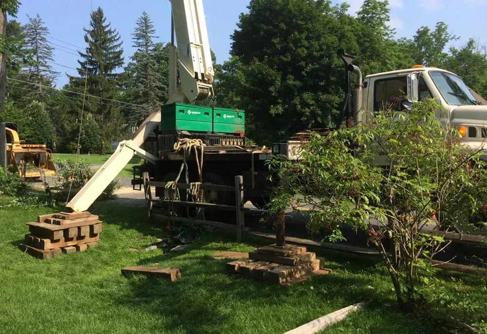 Truck with crane extended, on grass, near wooden fence. Green equipment boxes on truck bed.