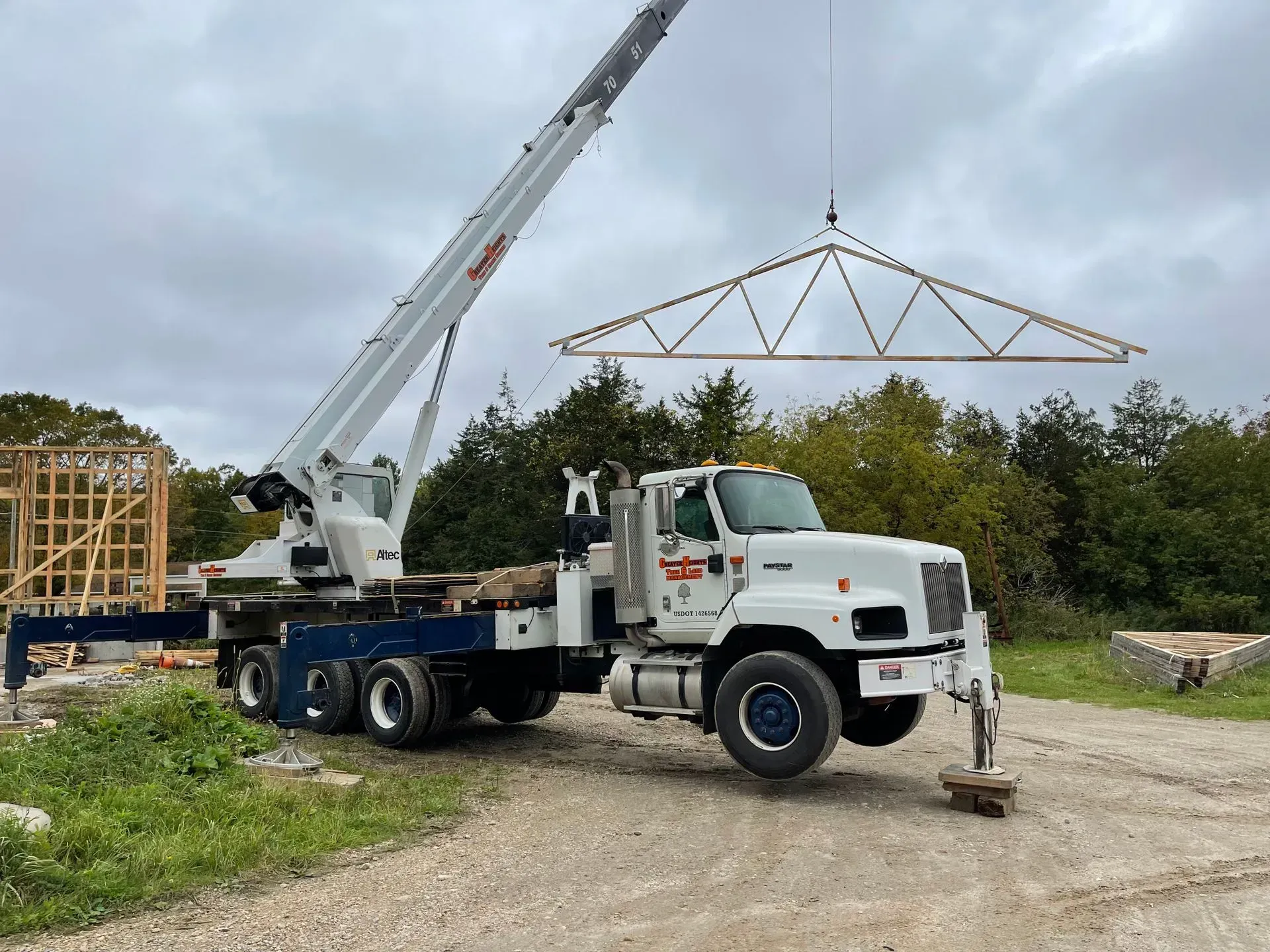 A white crane truck parked on a gravel site lifts a large wooden roof truss into the air against a cloudy sky.